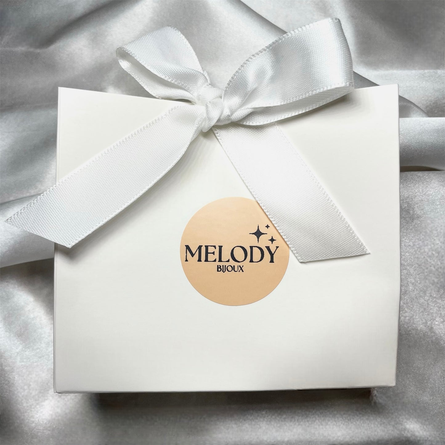 Petit sac cadeau élégant pour bijoux, en carton rigide avec nœud blanc, logo Melody Bijoux à l’avant et inscription dorée “Merci” au dos, parfait pour offrir avec style